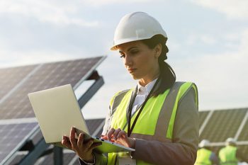 Portrait of female engineer in safety helmet and uniform using laptop checking solar panels.